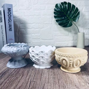 May include: Three decorative ceramic planters with different embossed designs. The planters are in shades of gray, white, and beige. The planters have a round base and a wide, round top.