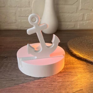 May include: White ceramic anchor phone stand with a round base. The anchor is a decorative element and the stand is designed to hold a phone upright.