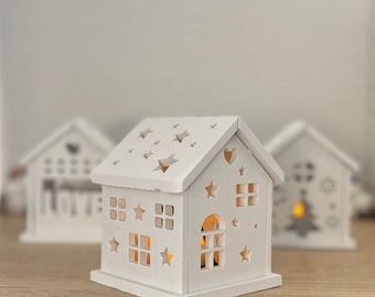 May include: Three white wooden houses with cut-out star and heart designs. The center house has a warm glow coming from inside.