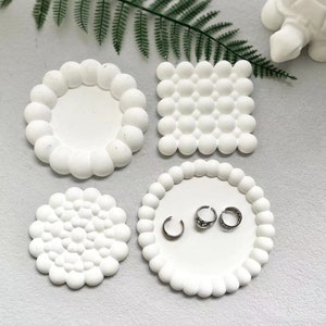 Peut inclure: Quatre coupelles à anneaux en céramique blanche avec des motifs texturés différents. Les coupelles sont rondes et ont un bord surélevé. Trois des coupelles sont vides et une coupelle contient trois anneaux argentés.