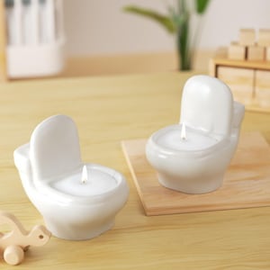 May include: Two white ceramic toilet-shaped candles with lit wicks. The candles are on a light wooden surface. One candle is on a small wooden board. A wooden toy turtle is in the lower left corner.