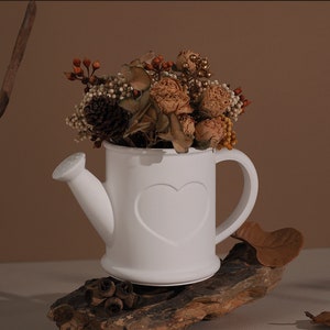 May include: White ceramic watering can with a heart design, filled with dried flowers and placed on a piece of driftwood.