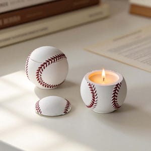 以下が含まれることがあります： 野球をテーマにした3つの装飾品：蓋付きの容器、火の灯ったキャンドルホルダー、小さな蓋。すべて白で、赤いステッチのディテールがあり、野球に似ています。キャンドルホルダーは温かい光を放っています。