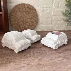 Puede incluir: Tres mini maceteros de cerámica blanca en forma de coche. Dos de los coches están vacíos y uno tiene pequeñas piedras rosas y moradas dentro.