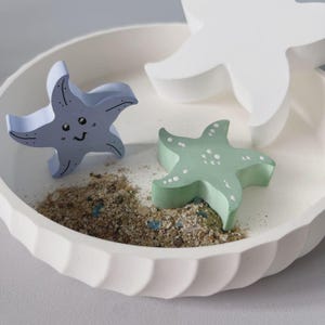 May include: A white, scalloped-edge bowl holds three starfish figurines and sand. Two starfish are painted light blue and green, with white accents. The third starfish is white. The bowl is on a light gray surface.