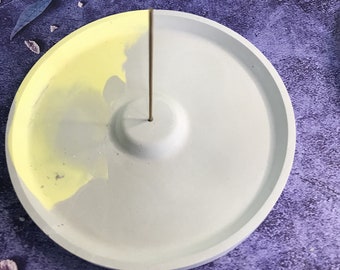 May include: A round, gray concrete incense holder with a yellow glaze on one side. The holder has a small, raised center for holding an incense stick.