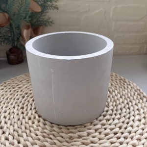 May include: A plain white cylindrical concrete planter pot, sitting on a woven wicker coaster.
