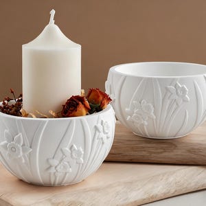 Op de afbeelding: Twee witte keramische kommen met reliëf narcisontwerpen. Eén kom bevat een grote witte kaars, gedroogde bloemen en decoratieve elementen. De andere kom is leeg. Beide kommen staan op een houten oppervlak, wat een serene, decoratieve presentatie creëert.