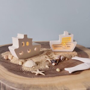 May include: Two off-white ceramic boat-shaped candle holders with cut-out windows and a warm glow. The candle holders are on a wooden surface with sand, seashells, and a starfish. The background is a light blue.