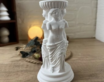 May include: White ceramic candle holder featuring three sculpted figures of women standing together. The figures are draped in flowing robes and have detailed facial features. The candle holder has a round base and a top opening for a candle.