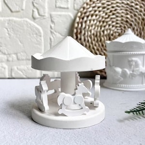 Könnte beinhalten: Ein weißes Miniatur-Karussell mit einem weißen Dach und Pferden. Das Karussell ist aus Keramik und hat ein schlichtes Design.