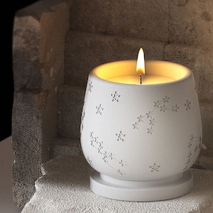 Pode incluir: Castiçal de cerâmica branco com um design de estrelas e uma vela acesa dentro. O castiçal tem uma base pequena e uma parte superior larga e arredondada.