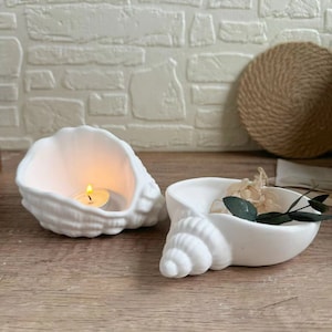 Op de afbeelding: Twee witte keramische schelpvormige kaarshouders. Een houder heeft een kleine theelichtkaars erin. De andere houder heeft gedroogde bloemen en groen erin.