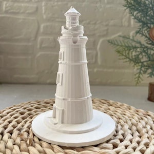 Puede incluir: Figurita de faro de cerámica blanca con un diseño detallado. El faro está sobre una base blanca y se encuentra sobre una superficie de mimbre tejido.