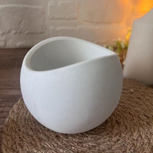 Può includere: Un vaso rotondo in ceramica bianca con un bordo liscio leggermente ondulato. Il vaso è appoggiato su una superficie intrecciata, marrone e testurizzata.