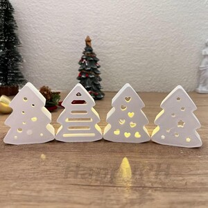 以下が含まれることがあります： ドット、水平線、ハート、星などの切り抜きデザインが施された、白いセラミック製のクリスマスツリーの装飾が4つ。木は内側から照らされ、温かい光を放っています。背景には他のクリスマスの装飾があります。