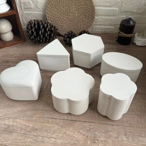 May include: Six white ceramic boxes in different geometric shapes, including a heart, a flower, a circle, a triangle, a hexagon, and another flower. The boxes are all made of the same material and have a smooth, matte finish.