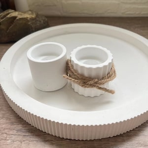 Op de afbeelding: Twee witte betonnen kaarshouders, een met een touw accent, op een witte ronde dienblad met een getextureerde rand.
