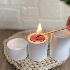 Può includere: Tre portacandele in ceramica bianca, uno con una candela tea light accesa. I portacandele sono su un tappetino di paglia intrecciata.