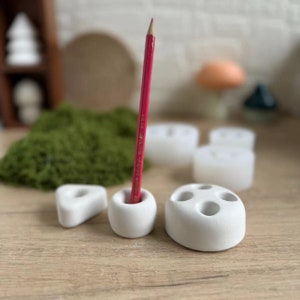 May include: White ceramic pencil holders of various sizes with a red pencil in one of the holders. The holders have multiple holes for pencils.
