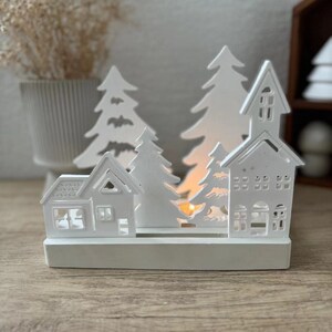 Puede incluir: Portavelas de madera blanca en forma de pueblo navideño con árboles y casas recortados. El portavelas tiene un espacio para una vela de té en el centro.