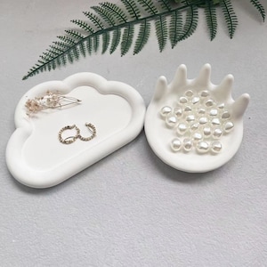 May include: Two white ceramic trays, one shaped like a cloud and the other like a hand. The hand tray holds a collection of white pearl beads.