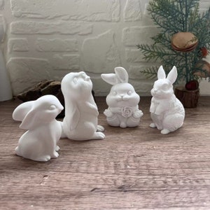 May include: Four white ceramic bunny figurines. The bunnies are in various poses, including sitting, standing, and looking up. The bunnies are all in a simple, minimalist style.