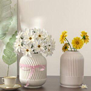 Può includere: Due vasi in ceramica bianca con fiori. Uno ha un nastro rosa e fiori bianchi con centri marroni. L'altro vaso ha fiori gialli. Una tazza e un piattino sono a sinistra.