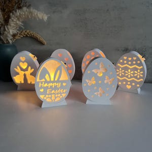 May include: Five white wooden Easter egg shaped light-up decorations. The eggs have different designs including bunnies, butterflies, hearts, and a zig-zag pattern. One egg says "Happy Easter".