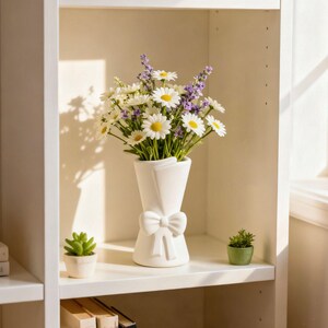 Op de afbeelding: Een witte keramische vaas in de vorm van een boeket, versierd met een strik, bevat een arrangement van madeliefjes en lavendel. Twee kleine potplanten staan ​​ernaast op een witte plank, wat een frisse uitstraling creëert.