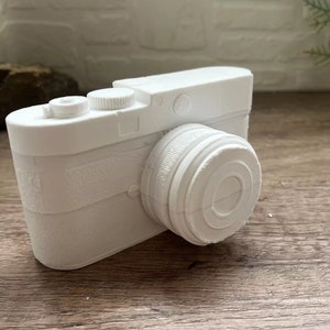 May include: A white ceramic camera figurine with a textured surface. The camera has a lens, a viewfinder, and a button on the top.