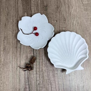 May include: Two white ceramic dishes: a flower shape with red berries and a seashell. The flower dish has petal details. The seashell dish has a textured, fan-like pattern. Both are on a wooden surface.