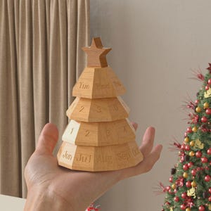 Può includere: Un calendario perpetuo a forma di albero di Natale in legno. Il calendario marrone chiaro presenta giorni, mesi e numeri incisi. L'albero ha una stella in cima ed è tenuto in una mano.