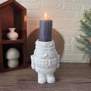 May include: White ceramic nutcracker candle holder with a gray candle inside. The candle is lit and the holder has a detailed design.