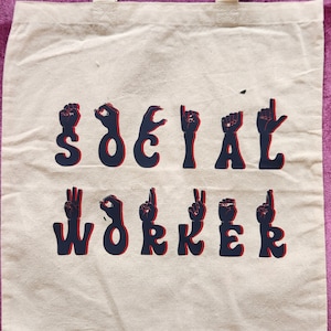 May include: Cream-colored tote bag with the words "SOCIAL WORKER" spelled out in American Sign Language. The letters are navy blue with a red shadow effect. The bag has two cream-colored handles.