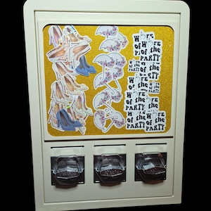 May include: A cream-colored vending machine display case with a gold glitter background. The display features stickers of high-heeled shoes, floral umbrellas, and the text "Wife of the Party" with a middle finger icon repeated multiple times.