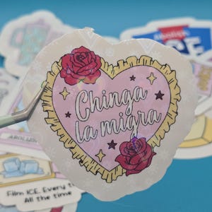 以下が含まれることがあります： 黒い筆記体で「Chinga la migra」と書かれたハート型のステッカー。赤いバラと星が描かれたピンク色のハートで囲まれています。ステッカーはピンセットで持たれており、背景には他のステッカーがあります。
