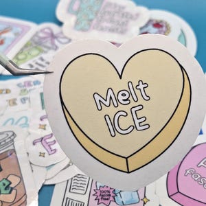 以下が含まれることがあります： 「Melt ICE」の文字が黒字で書かれた、黄色の縁取りのあるハート型のステッカー。ピンセットで持たれており、背景には他のステッカーが見える。