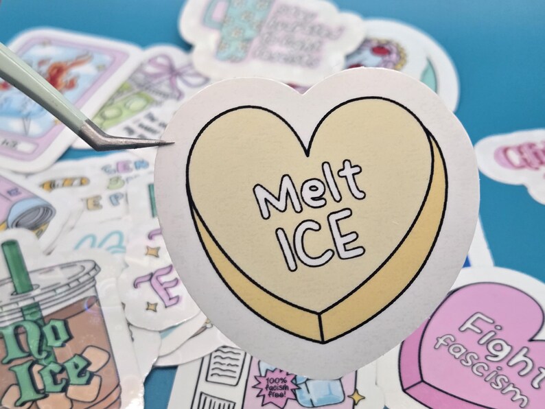 以下が含まれることがあります： 「Melt ICE」の文字が黒字で書かれた、黄色の縁取りのあるハート型のステッカー。ピンセットで持たれており、背景には他のステッカーが見える。