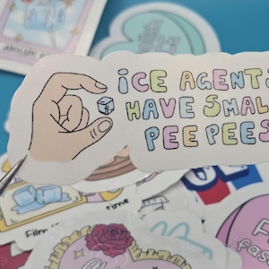 以下が含まれることがあります： パステルカラーで「ICE AGENTS HAVE SMALL PEE PEE'S」と書かれた白いステッカー。小さな氷のキューブを持っている手が表示されています。他のステッカーが背景に見えます。