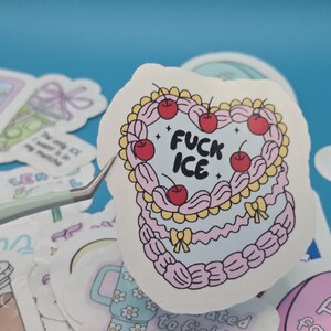以下が含まれることがあります： ピンク色のケーキのデザインで、さくらんぼと「FUCK ICE」の文字が描かれたハート型のステッカー。ステッカーは白い縁取りがあり、青い表面に他のステッカーに囲まれています。