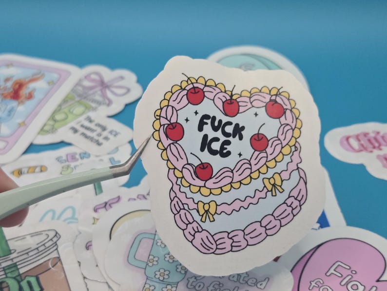 以下が含まれることがあります： ピンク色のケーキのデザインで、さくらんぼと「FUCK ICE」の文字が描かれたハート型のステッカー。ステッカーは白い縁取りがあり、青い表面に他のステッカーに囲まれています。