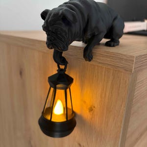 Könnte beinhalten: Eine schwarze Bulldoggen-Figur auf einer Holzoberfläche, die eine schwarze Laterne mit warmem LED-Licht hält. Die Laterne hängt an der Pfote des Hundes und bildet ein einzigartiges Dekorationsobjekt.