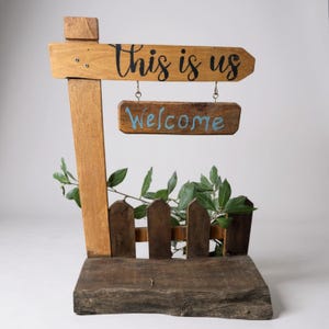 Puede incluir: Cartel de madera rústico con las palabras "This is us" y "Welcome". El cartel presenta una pequeña valla de madera y vegetación. El cartel es de madera y tiene un aspecto envejecido.