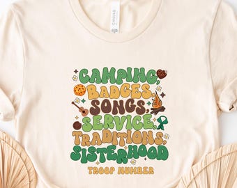 May include: A cream-colored t-shirt featuring a graphic with the text CAMPING, BADGES, SONGS, SERVICE, TRADITIONS, SISTERHOOD, and TROOP NUMBER. The design includes illustrations of a cookie, a guitar, a campfire, and a green ribbon.