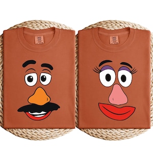 May include: Two brown t-shirts with cartoon faces, one with a mustache and orange nose, the other with eyelashes and a pink nose. The shirts are on a woven mat. Each shirt has a tag that says "COMFY COLORS".