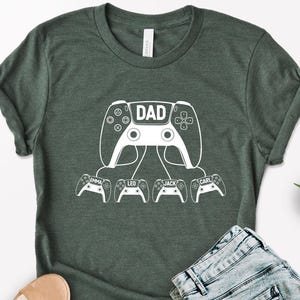 May include: Dark green t-shirt with a white graphic of a video game controller with the word "DAD" in the center. Below are smaller controllers with the names "EMMA", "LEO", "JACK", and "CARL".