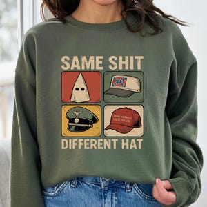 May include: Olive green sweatshirt with the text "SAME SHIT DIFFERENT HAT". The graphic features four images of different hats, including a KKK hood, a Confederate flag hat, a military hat, and a "Make America Great Again" hat.