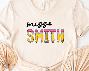 May include: Cream-colored t-shirt featuring the text "miss Smith" in a playful design. The word "miss" is in black script, while "Smith" is designed to look like pencils with yellow, pink, and black accents. A casual and fun teacher's tee.