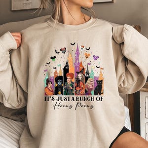 May include: Beige sweatshirt featuring a colorful graphic of the Sanderson sisters from Hocus Pocus, with a castle backdrop and bats. The text "IT'S JUST A BUNCH OF Hocus Pocus" is printed below the graphic.